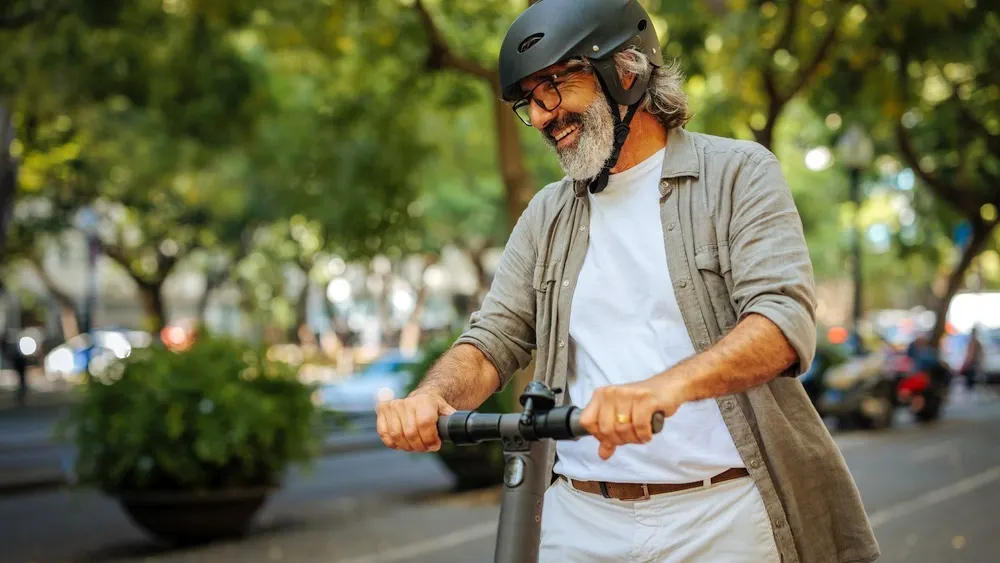 Ein Mann mit Brille und Helm fährt einen Roller auf einer Straße mit Bäumen im Hintergrund.