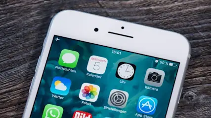 Nahaufnahme eines weißen Smartphones mit Apps auf einem Tisch. Apps wie Nachrichten, Kalender, Uhr und Kamera sind zu sehen.