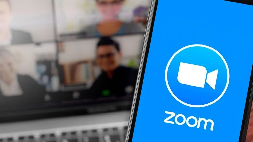 Blaues Handy mit Zoom-Logo im Vordergrund, unscharfer Bildschirm mit Personen im Hintergrund.