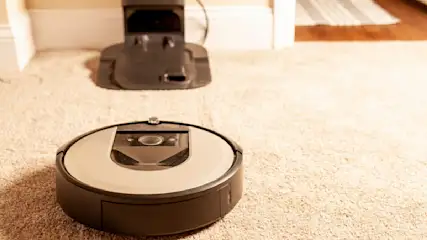 Ein runder Staubsaugerroboter steht auf einem beigen Teppich, mit einer Ladestation im Hintergrund vor einer weissen Wand.
