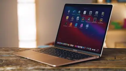 Laptop auf Holztisch mit bunten App-Symbolen auf dem Bildschirm. Hintergrund: weisse Wand und ein Hauch von Grün.