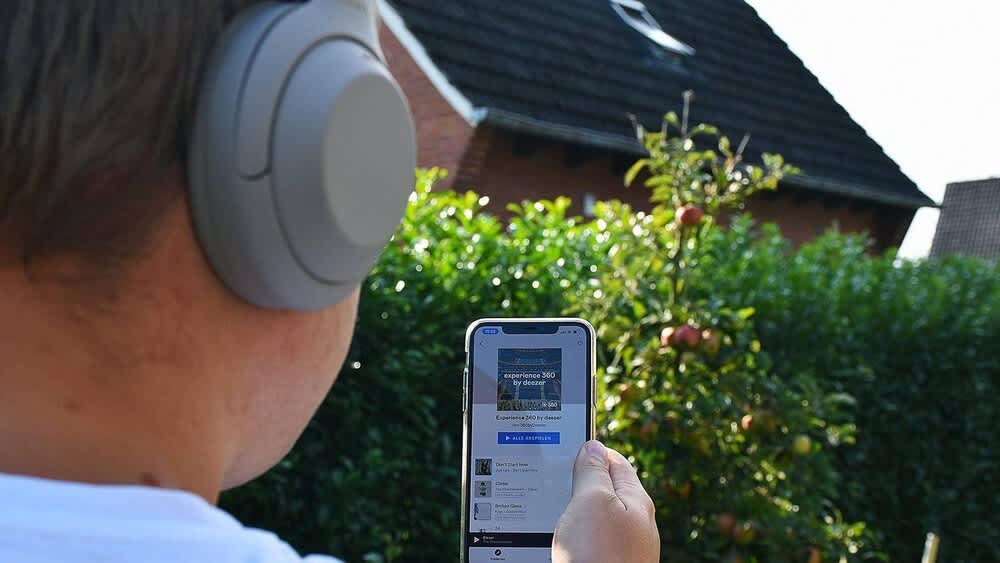 Alexander Mundt mit Over-Ear-Kopfhörern Sony WH-1000XM4 von hinten zu sehen. Er hält ein Smartphone in der Hand, auf der eine App geöffnet ist.