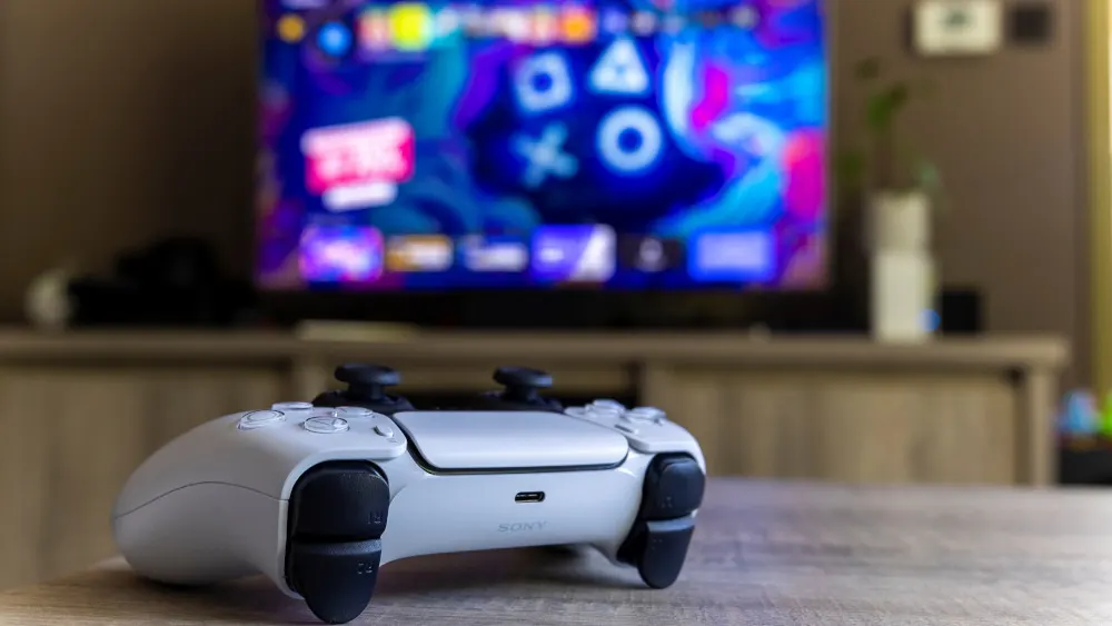 Ein weißer PlayStation 5-Controller liegt auf einem Holztisch. Im Hintergrund ist ein eingeschalteter Fernseher auf einem Holzschrank zu sehen.