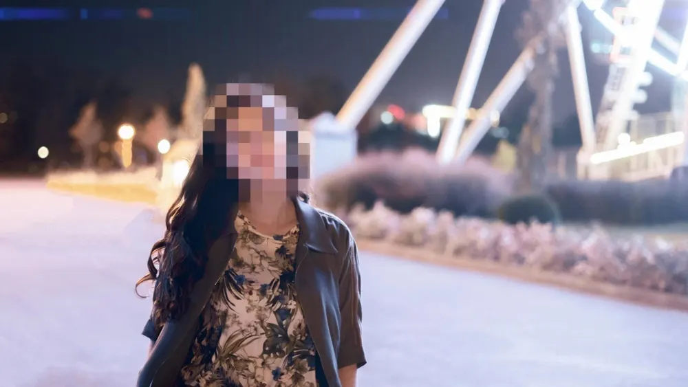 Eine Person mit gemustertem Oberteil und Jacke steht vor einem beleuchteten Riesenrad im Freien.