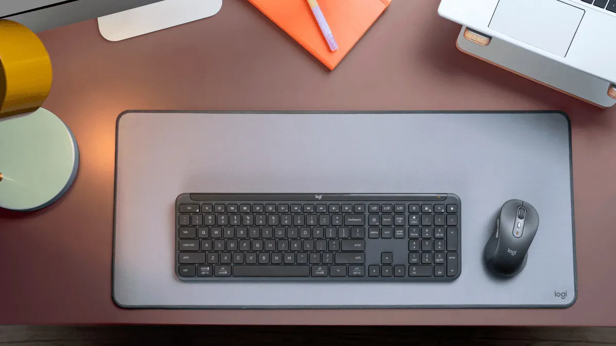 Ein Schreibtisch mit einer grauen Matte, einer schwarzen Tastatur, einer Maus und einem Notizbuch. Ein Laptop ist im Hintergrund.