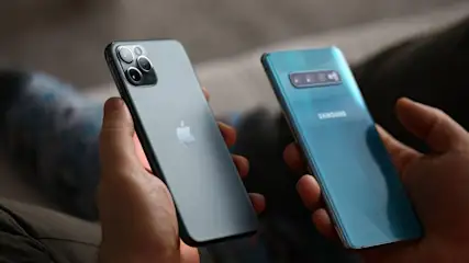 Zwei Hände halten ein graues iPhone mit drei Kameras und ein blaues Samsung-Handy mit drei Kameras auf der Rückseite.