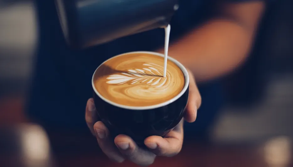 Milch wird in einen schwarzen Becher mit Latte Art gegossen, der in den Händen einer Person gehalten wird.