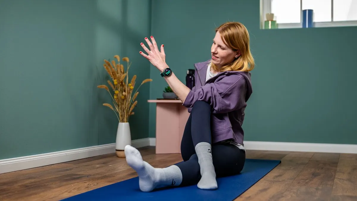 Frau sitzt auf einer blauen Yogamatte, trägt eine lila Jacke, schwarze Leggings und graue Socken. Sie streckt eine Hand aus.