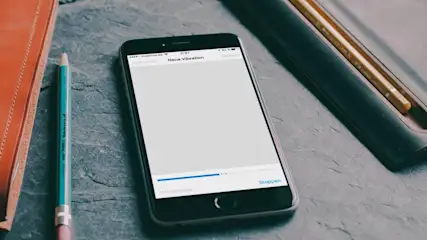 Auf einem Tisch liegen ein Handy mit weißem Bildschirm, ein orangefarbenes Notizbuch, ein blauer Stift und ein Stiftehalter.
