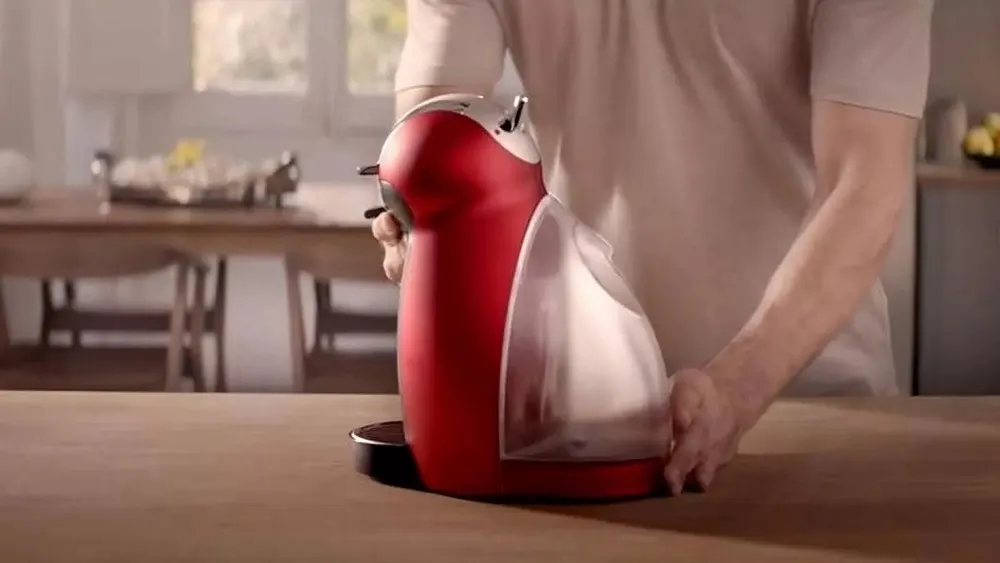 Eine rote Kaffeemaschine steht auf einem Holztisch, der mit Händen bedient wird. Im Hintergrund befinden sich ein Tisch und Schränke.