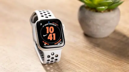 Eine Smartwatch mit weißem Locharmband zeigt auf dem Display die Uhrzeit 10:41. Im Hintergrund steht eine grüne Topfpflanze auf einem Holztisch.