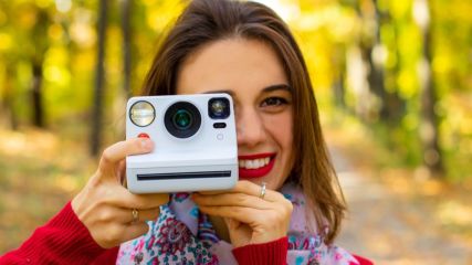 Eine lächelnde Frau hält eine Polaroid Kamera vor ihr Gesicht 