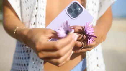 Eine Person hält ein lila Handy und zwei lila Gänseblümchen vor einem Strand. Die Person trägt eine weiße Häkeljacke.
