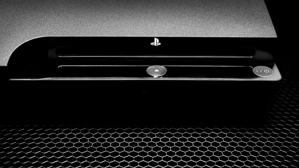 Eine graue Playstation 3 Konsole ist auf einer schwarzen, sechseckigen Oberfläche platziert. Das Playstation-Logo ist auf der Konsole abgebildet.