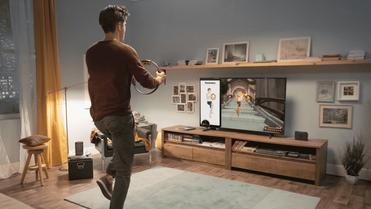 Junger Mann steht im Wohnzimmer vor dem Fernseher mit Ring-Con in der Hand und spielt Ring Fit Adventure