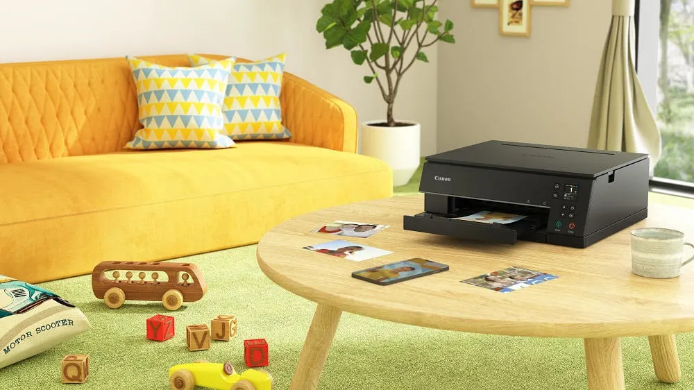 Gelbes Sofa mit Kissen, Tisch mit Drucker, Fotos und Tasse. Spielzeug auf grünem Teppich. Zimmerpflanze im Hintergrund.