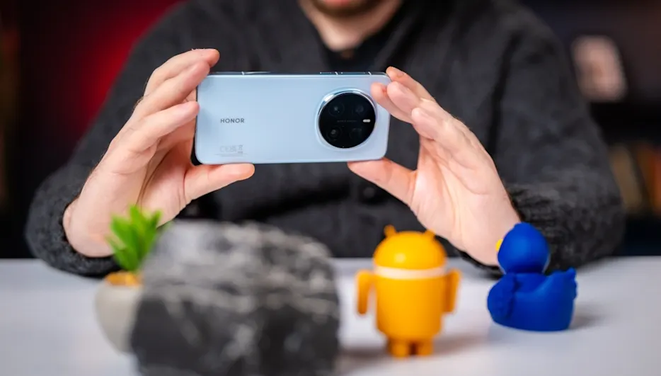 Mann hält blaues Honor-Handy. Tisch mit Pflanze, Stein, gelber Android-Figur und blauer Ente.
