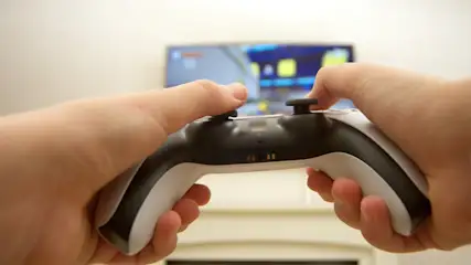 Zwei Hände halten einen schwarz-weißen Controller vor einem unscharfen Fernseher und einem hellen Hintergrund.