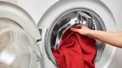 Eine Hand legt einen roten Pullover in eine offene, weiße Waschmaschine.