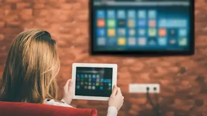 Frau hält Tablet vor roter Ziegelwand. Fernseher und Steckdose sind ebenfalls sichtbar.