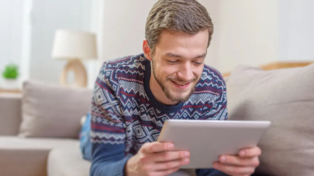 Ein Mann im gemusterten Pullover liegt auf einer Couch und benutzt ein Tablet. Im Hintergrund sind eine Lampe und Kissen zu sehen.