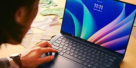 Eine Person tippt auf einem Laptop mit blauem Hintergrund und der Uhrzeit 11:11. Daneben liegen Stoffreste und eine Armbanduhr.