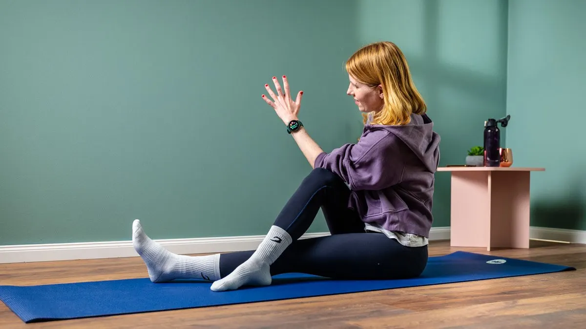 Eine Person sitzt auf einer blauen Yogamatte, trägt Socken, Leggings und einen lila Kapuzenpullover. Im Hintergrund sind eine grüne Wand und ein Tisch.