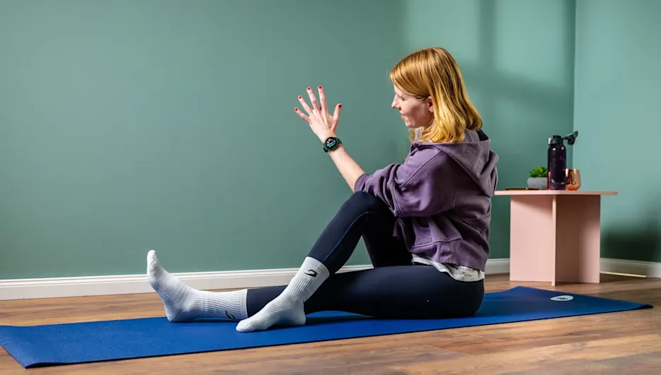 Eine Person sitzt auf einer blauen Yogamatte, trägt Socken, Leggings und einen lila Kapuzenpullover. Im Hintergrund sind eine grüne Wand und ein Tisch.