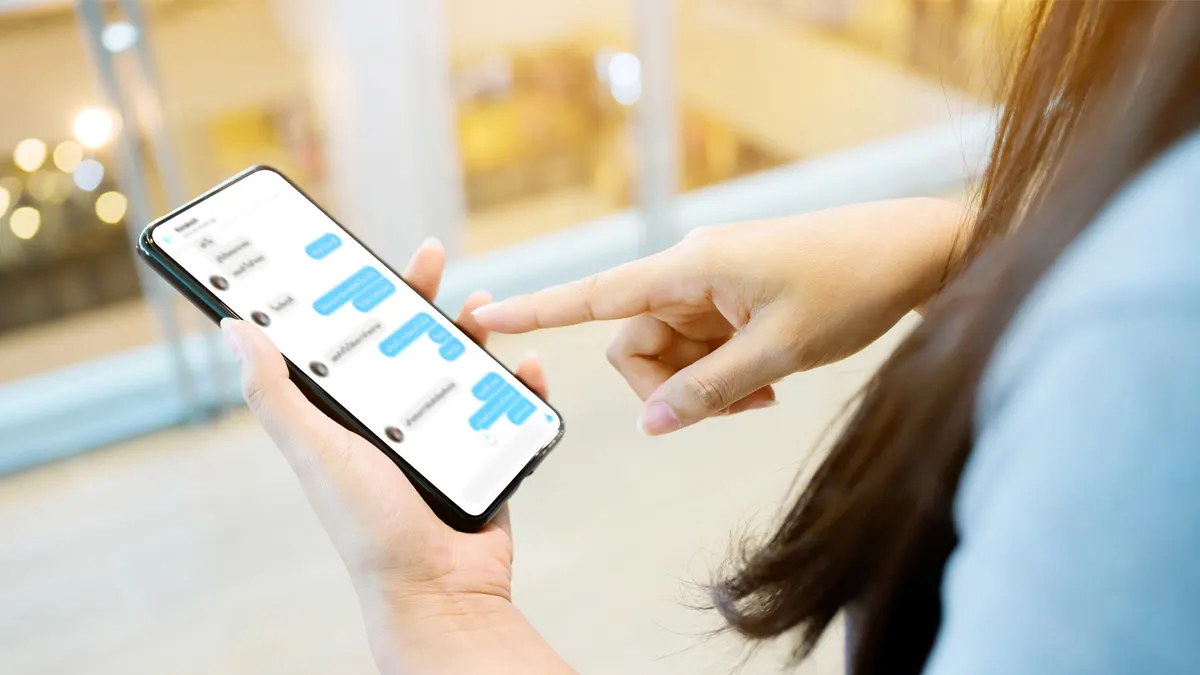 Eins Smartphone liegt in der Hand einer Frau, die mit der anderen Hand darauf tippt. Es ist ein unscharfer Chatverlauf erkennbar.