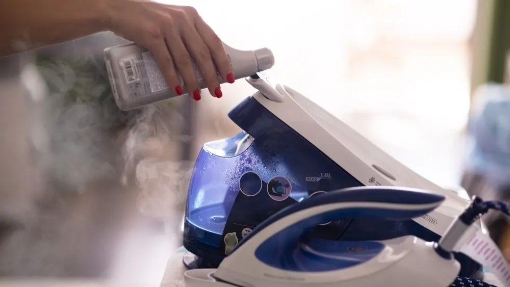 Eine Hand gießt Flüssigkeit aus einer Flasche in ein Dampfbügeleisen mit blauem Wassertank, Dampf steigt auf.