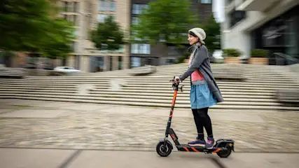Eine Frau fährt auf einem orangefarbenen Elektroroller durch eine städtische Umgebung mit Gebäuden und Treppen.