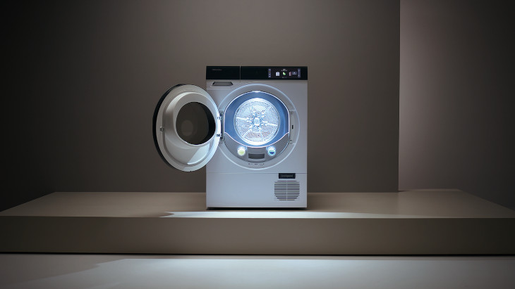 Ein Wäschetrockner mit geöffneter Tür steht auf einem Podest vor einem grauen Hintergrund. Die Trommel ist blau beleuchtet.