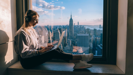 Eine Frau trägt Kopfhörer und sitzt auf einer Fensterbank mit einem Laptop. Im Hintergrund eine Skyline