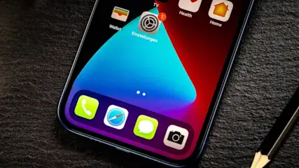 Nahaufnahme eines Smartphones mit Apps auf dem Bildschirm, neben einem Bleistift auf dunklem Grund. Apps wie Wallet, Einstellungen und Home sind sichtbar.
