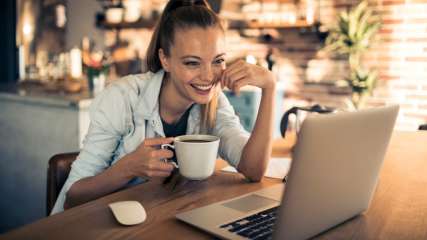 Eine lächelnde Frau sitzt vor einem Laptop in einem Cafe.