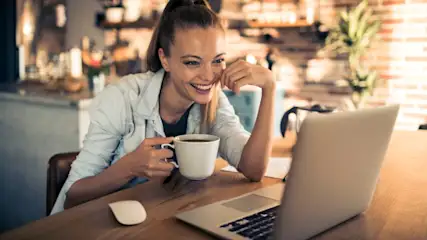 Eine Frau sitzt an einem Holztisch mit einem Laptop und einer Tasse. Im Hintergrund sind Regale und eine Ziegelwand zu sehen.