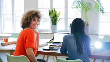 Zwei Frauen sitzen an einem Tisch mit Laptop und Tablet. Eine Frau lächelt und trägt einen orangefarbenen Pullover. Pflanzen im Hintergrund.