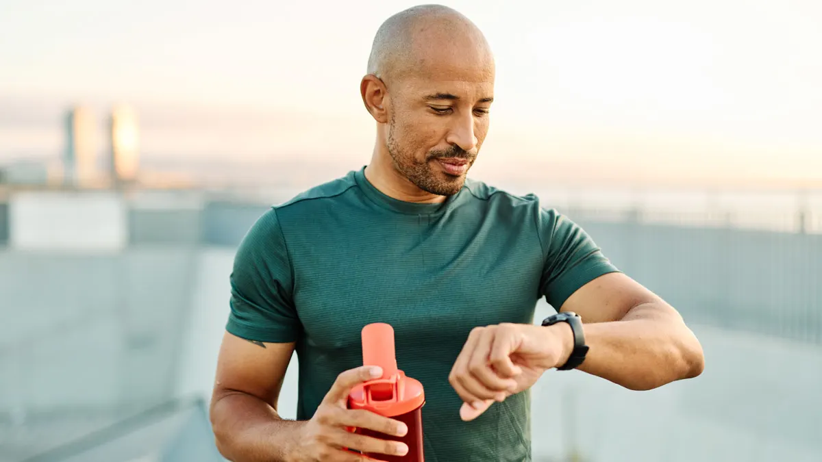 Ein muskulöser Mann steht mit Sporttrinkflasche frontal zur Kamera und blickt auf seine Smartwatch am Handgelenk.