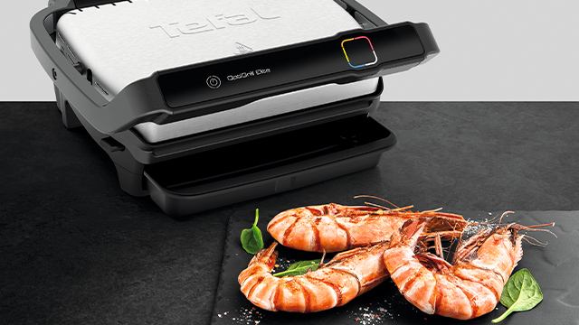 Ein schwarzer Grill steht neben gegrillten, orangefarbenen Garnelen und grünen Blättern auf einer schwarzen Oberfläche.