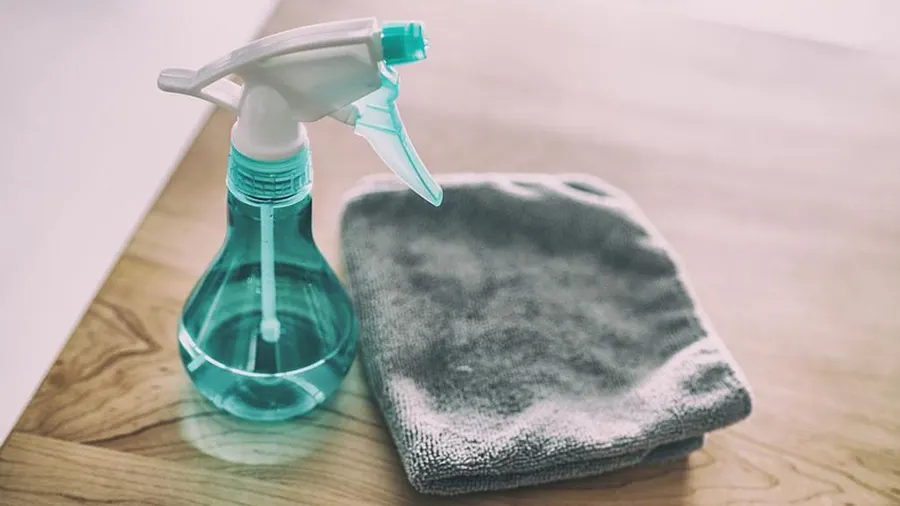 Ein türkisfarbener Sprühflasche und ein graues Tuch liegen auf einer Holzoberfläche vor einer weißen Wand.
