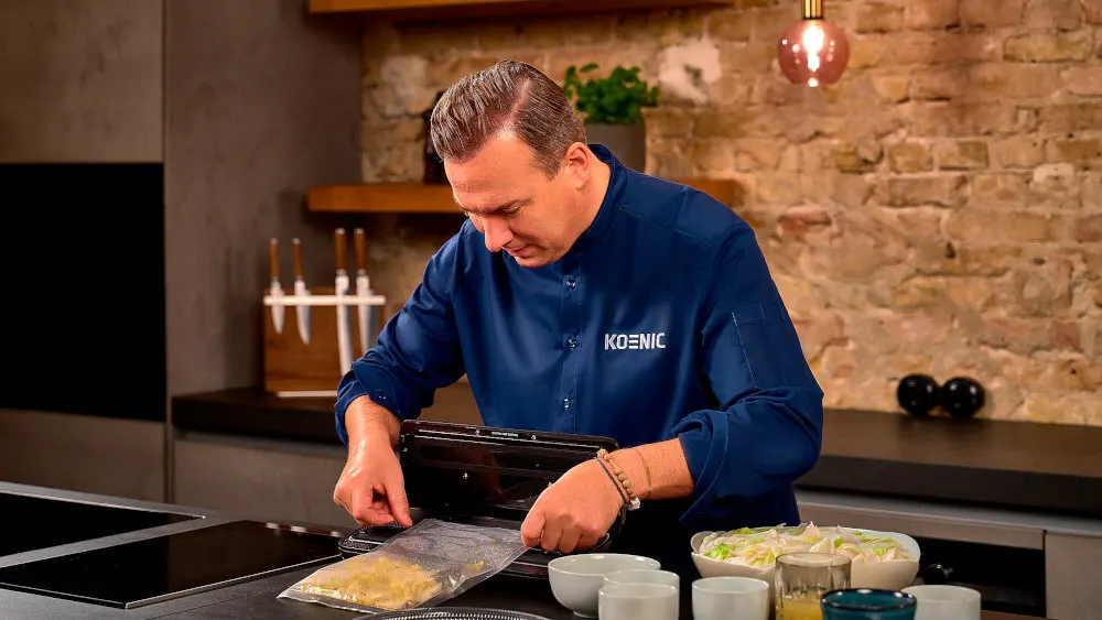 Ein Mann mit blauem Hemd vakuumiert eine Tüte mit Essen in einer Küche, Schüsseln und Beilagen sind auf der Arbeitsfläche.