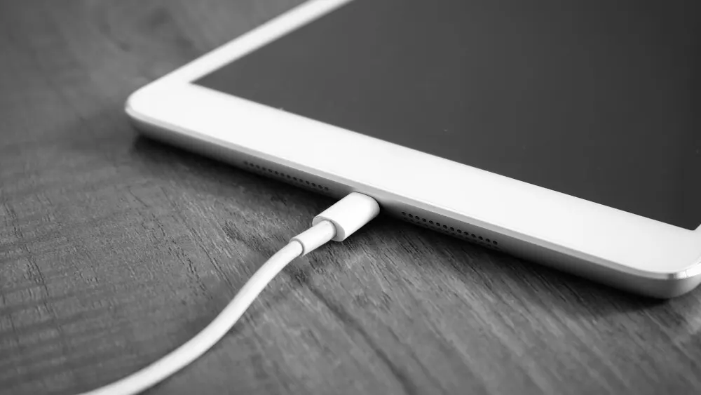 Ein Tablet mit schwarzem Bildschirm liegt auf einem Holztisch. Ein weißes Ladekabel ist an das Gerät angeschlossen.