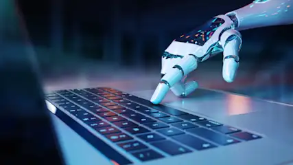Eine Roboterhand tippt auf einer Laptop-Tastatur. Der Hintergrund ist unscharf und dunkel. Die Tasten sind rechteckig und schwarz.