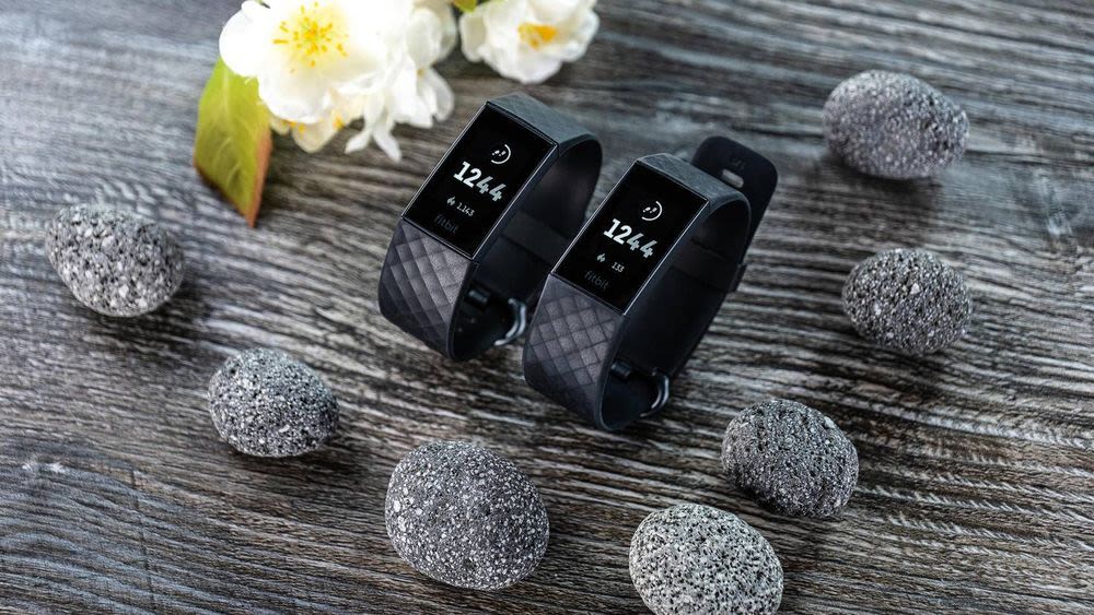 Fitbit Charge 4 und Fitbit Charge 3 auf Holzoberfläche mit Dekotsteinen und -blumen