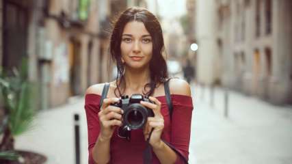 Portrait einer Fotografin, die mit Kamera in beiden Händen in einer Gasse steht.