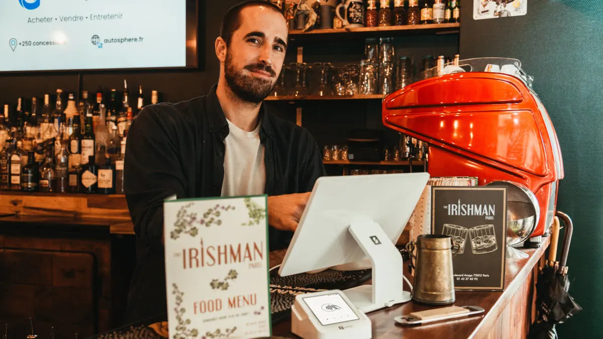 Ein Mann steht an einer Bar mit Getränken, einem Menü, einem weißen Tablet und einer roten Kaffeemaschine.