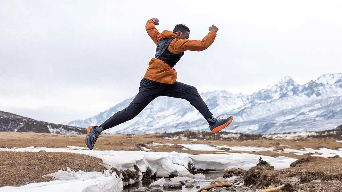 Mann in Sportkleidung springt über einen Bach mit schneebedeckten Bergen im Hintergrund. Er trägt eine orangefarbene Jacke und dunkle Hosen.