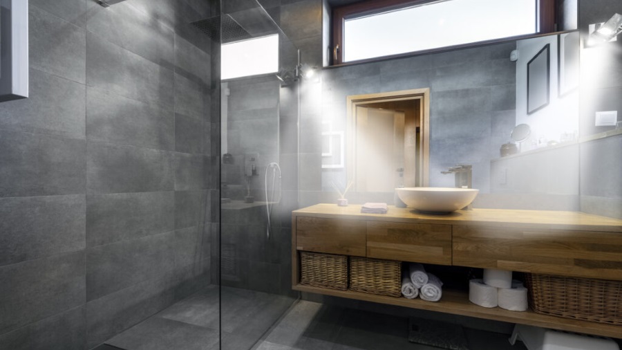 Ein Badezimmer mit grauen Fliesen, einem Holztisch mit Waschbecken und Körben. Handtücher und Toilettenpapier sind darunter verstaut.