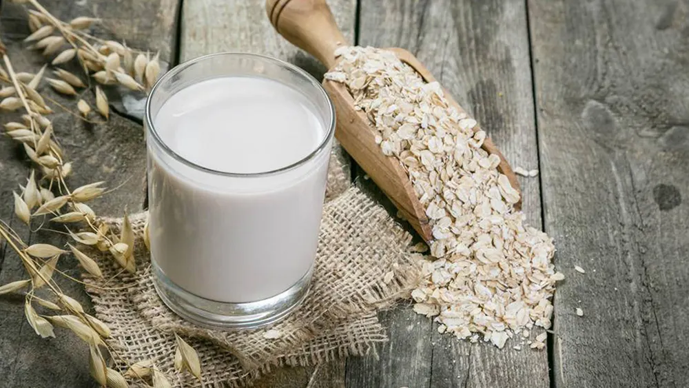 Auf einem Tisch stehen ein Glas Hafermilch, ein Holzschaufel mit Haferflocken und ein Jutesack.