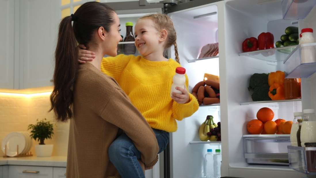 Eine Frau hält ein Kind auf den armen und steht vor einem Kühlschrank.